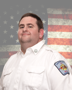 A person in a white uniform shirt with a badge on the sleeve stands confidently. They are set against a backdrop of a weathered American flag.