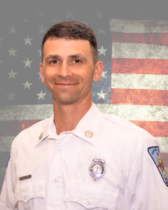 A man in a white uniform shirt with buttons and patches smiles against a blurred American flag backdrop.