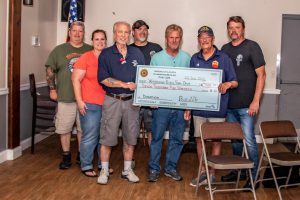 A group of seven people, including several men and women, stand together indoors holding a large novelty check. One person wears a shirt with an American flag logo. An American flag stands in the background. They appear to be celebrating a donation.