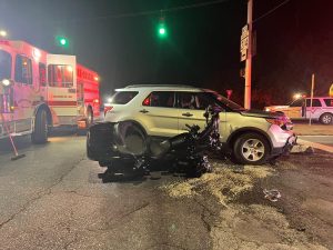 A car and a motorcycle involved in a collision at an intersection. The motorcycle is on its side, with debris around. An emergency vehicle is present, and the traffic light is green. The scene is lit by streetlights and emergency lights.