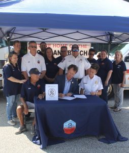 A group of people under a canopy. Two individuals in the foreground are seated at a table, one signing a document. Others stand around them, some in uniform. A vehicle with "Home of the St. Andrews Beach" is partially visible in the background.