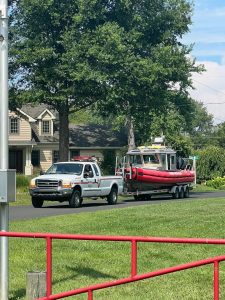 A white truck is towing a red rescue boat on a trailer down a suburban street. The street is lined with trees and houses. In the foreground, there's a grassy area with a red fence. The sky is partly cloudy.