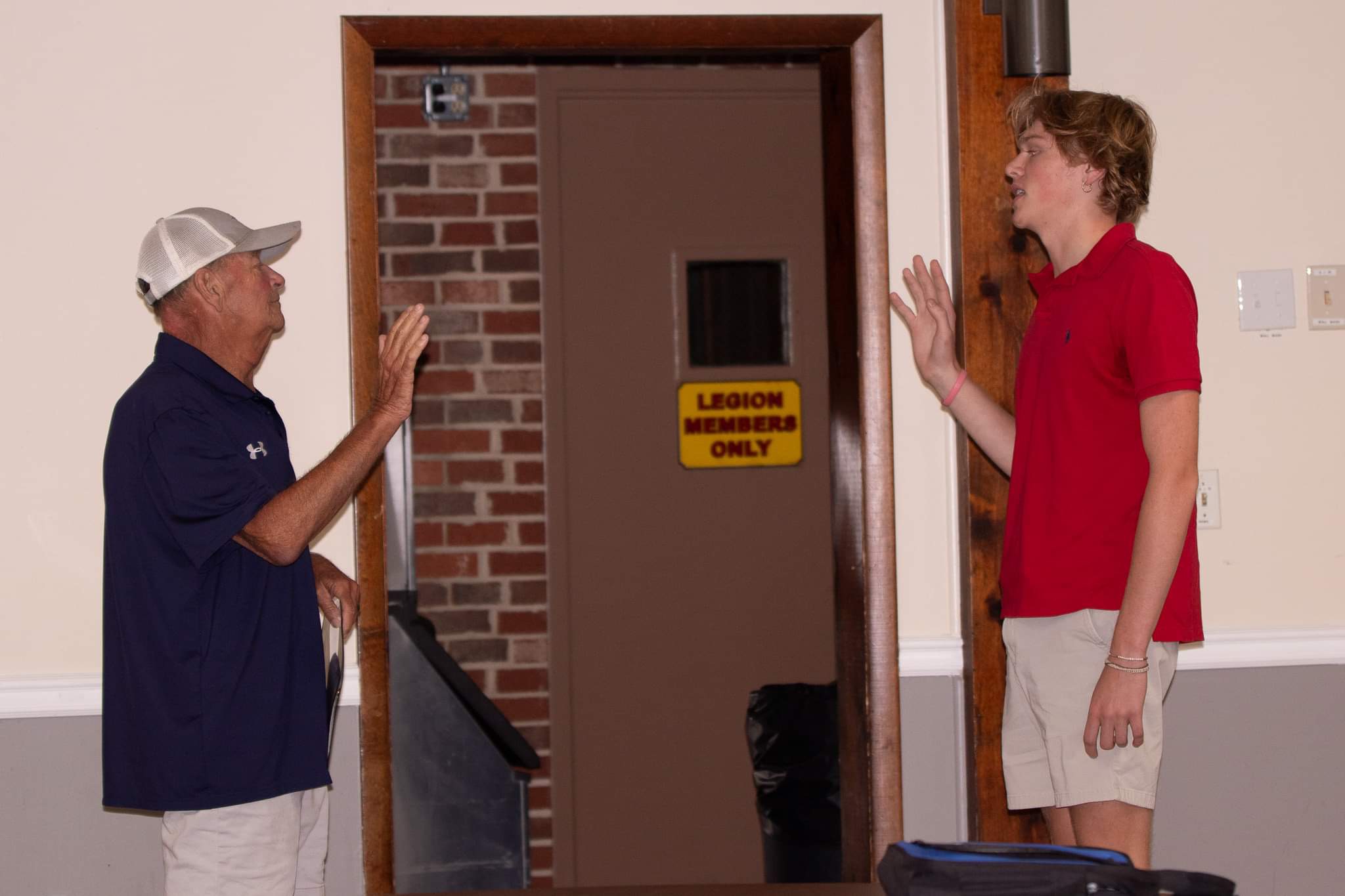 Two people in casual attire stand facing each other, raising their hands as if in greeting or conversation. The older person wears a cap and a navy shirt, while the younger person wears a red shirt. A door in the background has a "Legion Members Only" sign.