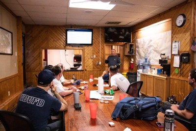 A group of firefighters sit around a wooden table in a meeting room. They are watching a presentation on a TV screen, given by a person standing at the front of the room. The room has wooden walls, a map, and various equipment.