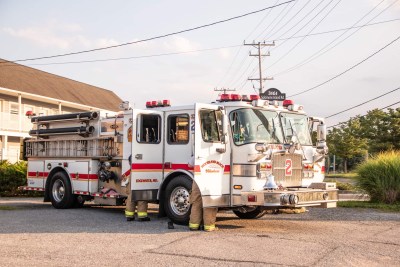 A fire truck with two firefighters standing beside it is parked on a paved area. The truck is white with red stripes and marked with the number 2. Nearby are a building and utility poles, under a partly cloudy sky.