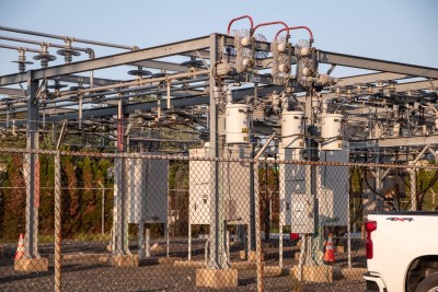 A fenced electrical substation with transformers and wiring. The structure has metal frames and various components. Part of a white pickup truck is visible in the lower right corner. Trees and a clear sky are in the background.