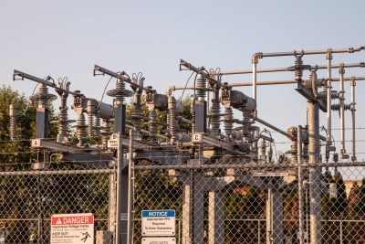 Electrical substation equipment surrounded by a chain-link fence. Warning signs on the fence indicate danger and required protective gear. The substation features transformers, insulators, and various metal components under a clear sky.