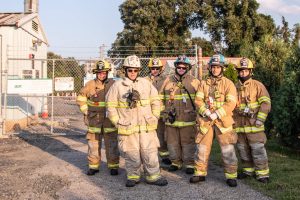 Six firefighters in full gear stand together outside a fenced facility with signs. They are smiling, and the background includes trees and a clear sky.