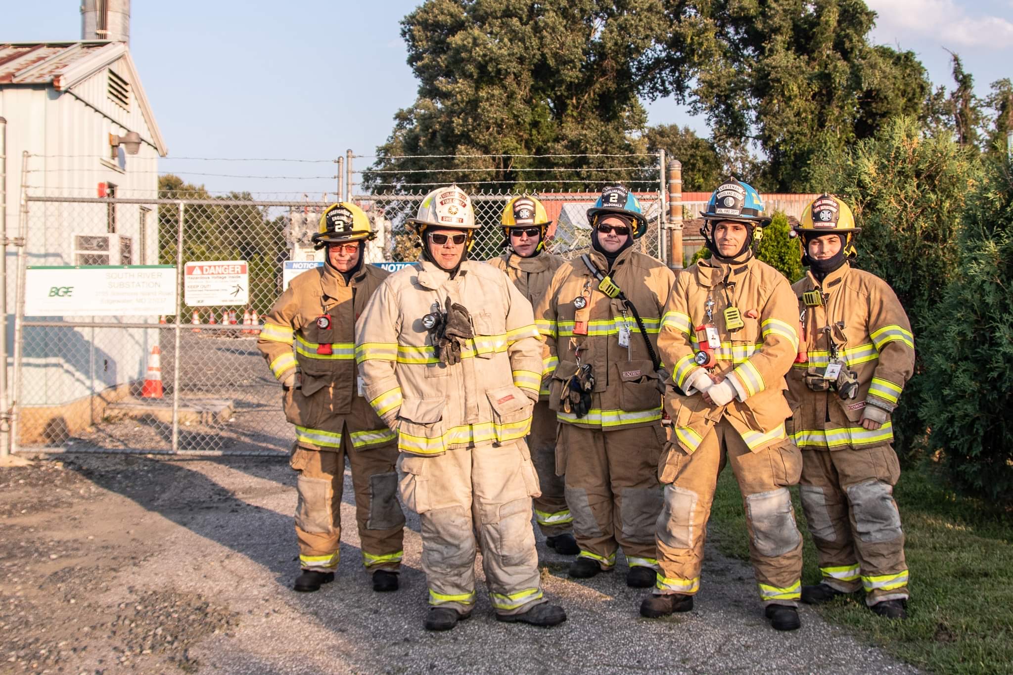 Six firefighters in full gear stand together outside a fenced facility with signs. They are smiling, and the background includes trees and a clear sky.