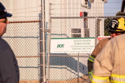 Workers stand near a chain-link fence outside the South River Substation in Edgewater, MD. A sign displays the address and a hazard hotline number. One worker wears a helmet marked with "2.