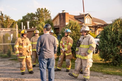 A group of firefighters in gear stand and listen to a person wearing a blue helmet and gray long-sleeve shirt. They are outdoors near a chain-link fence and a brick house, with trees and a clear sky in the background.