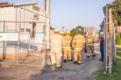 A group of firefighters in full gear walk away from the camera towards a building at a substation. They are passing through a gate with warning signs. Trees and a utility structure are visible in the background. The scene is well-lit by daylight.