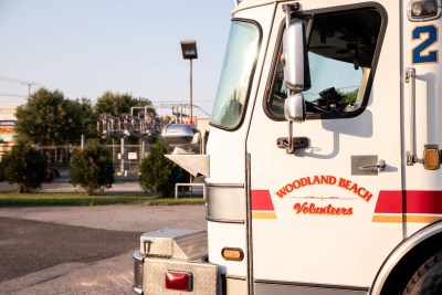 Side view of a fire truck labeled "Woodland Beach Volunteers" parked in a lot. Background includes trees, a utility pole, and some buildings under a clear sky.
