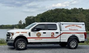 A white Woodland Beach Volunteer Fire Rescue truck is parked on a gravel path beside a body of water. The truck has red and yellow lettering with a fire department emblem. Trees and a cloudy sky are in the background.