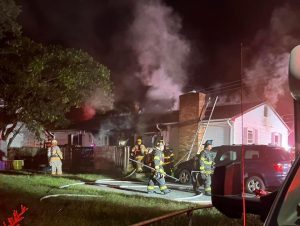 Firefighters work to extinguish a house fire at night. Smoke billows from the roof of a one-story house. Several firefighters in gear manage hoses, while emergency lights illuminate the scene. A car is parked near the house.