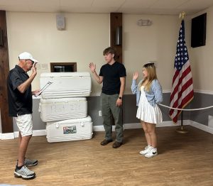 A man in a black shirt and cap stands with a paper, facing a young man and woman with raised right hands in a room with a wooden floor. Behind them are stacked coolers labeled "Post 226" and a U.S. flag by the wall.