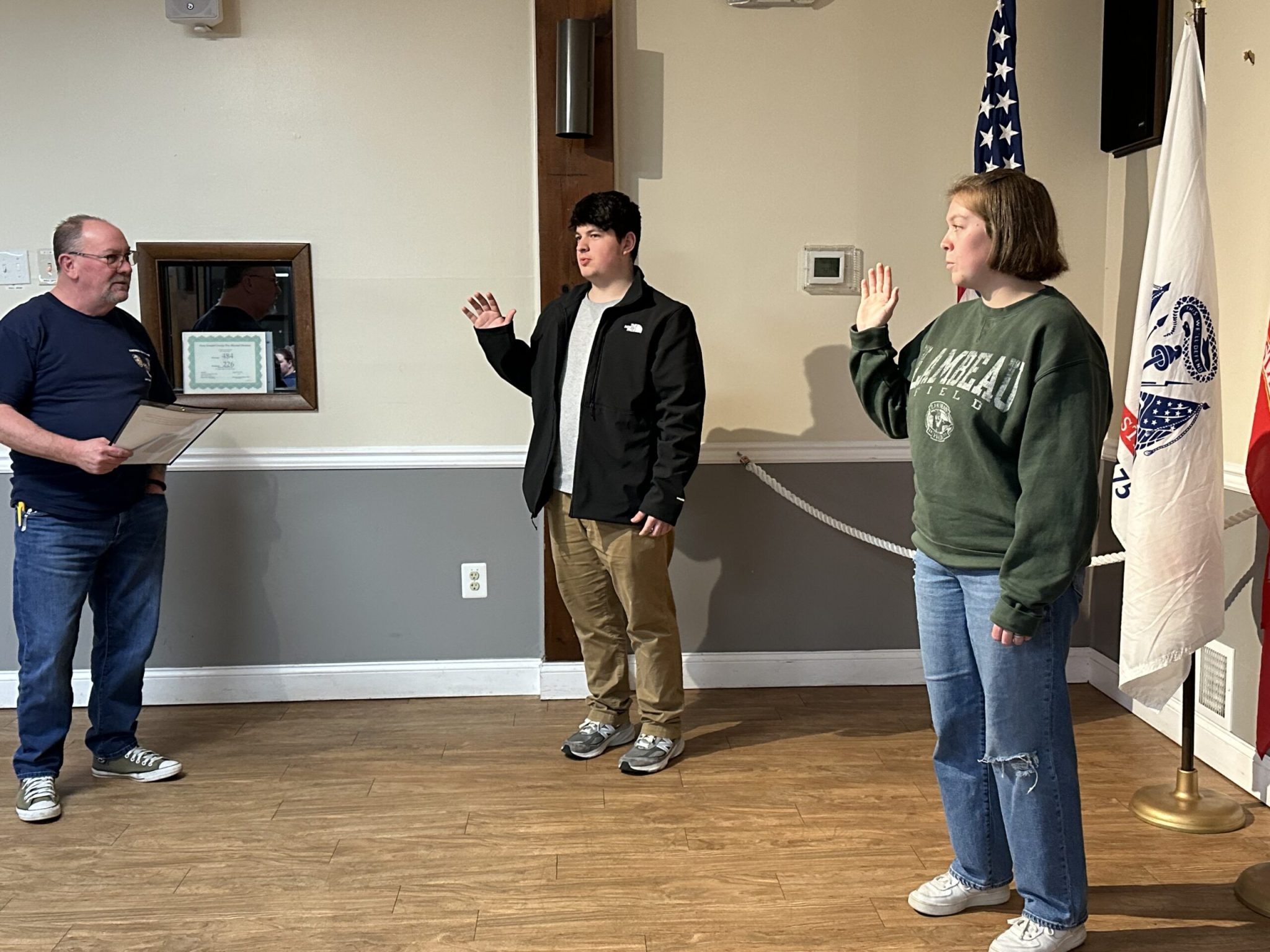 Three people in a room: one holding a paper, two with raised right hands as if taking an oath. A certificate is visible on the wall, and there are flags in the background. The floor is wooden.