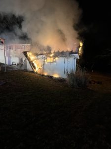 A small house engulfed in flames at night, with thick smoke rising into the dark sky. The fire is concentrated on the roof and side of the structure. The surrounding area is grassy and dimly lit.