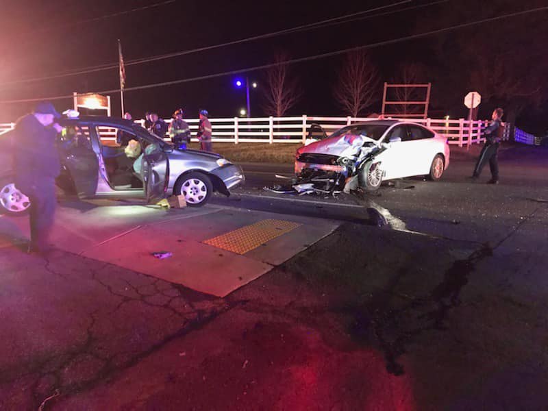 Two cars, one silver and one white, are involved in a nighttime collision at an intersection. The silver car's front side and white car's front are damaged. Emergency responders and a police officer are on the scene. A wooden fence is visible in the background.