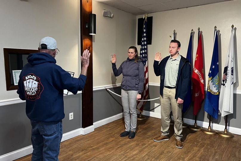 Three people in a room with one administering an oath to a woman and man standing with raised hands. Behind them are five flags, including the American flag, on stands. The room has wooden floors and a spotlight on the wall.