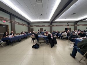 A large group of people sits around tables arranged in a U-shape in a community hall. Some are speaking while others listen attentively. The room is well-lit, with beige walls and a tiled floor. Casual attire suggests an informal meeting.