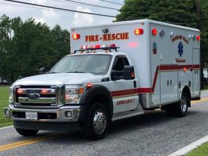 A white and red fire-rescue ambulance is driving on a road, with emergency lights on. The background features trees and a clear sky. The vehicle is marked with "Pocomoke City Volunteer" and "Ambulance 2.