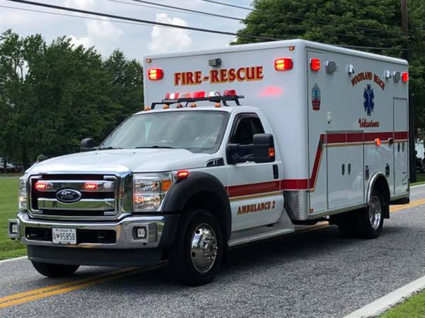 A white and red fire-rescue ambulance is driving on a road, with emergency lights on. The background features trees and a clear sky. The vehicle is marked with "Pocomoke City Volunteer" and "Ambulance 2.