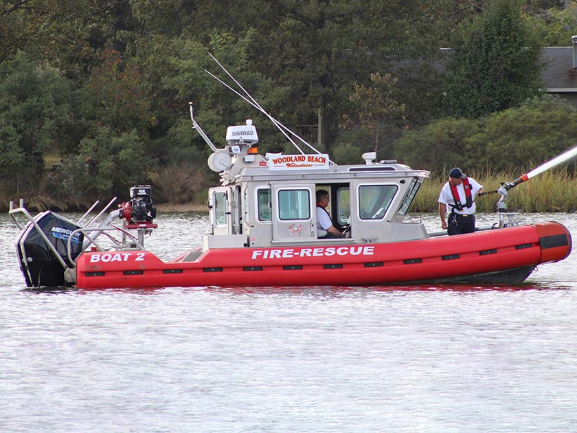 A red and white fire-rescue boat labeled "Boat 2" is stationed on a body of water. One person is visible operating a water hose. The background has trees and grass.