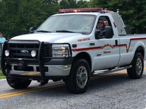 A white Ford fire-rescue truck with red stripes is parked on a road. The truck bears the words "Woodland Hills Volunteer Fire-Rescue." A person is visible in the driver's seat. Trees are in the background.