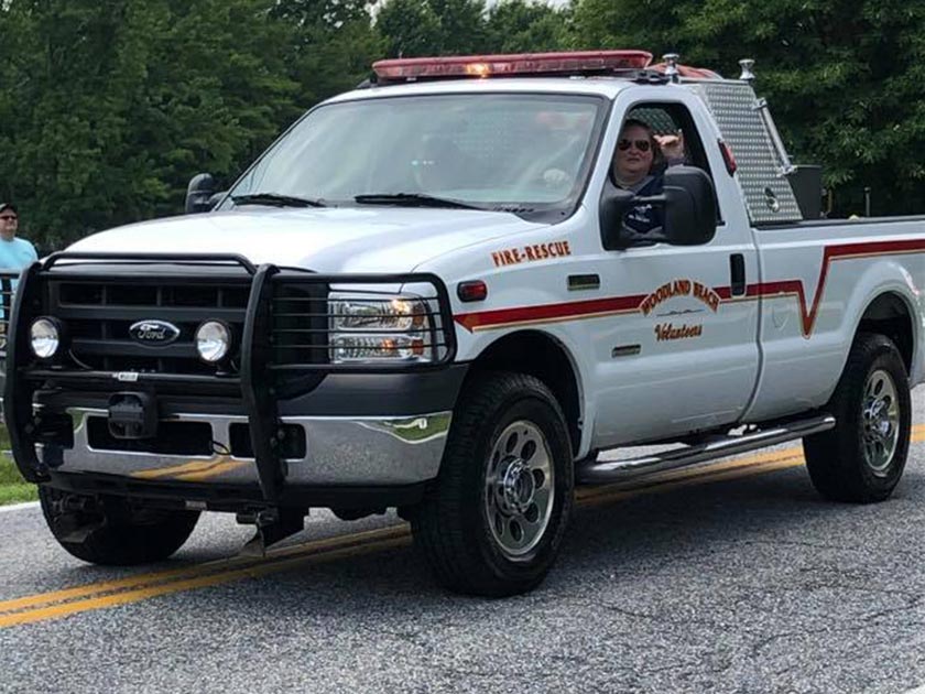 A white Ford fire-rescue truck with red stripes is parked on a road. The truck bears the words "Woodland Hills Volunteer Fire-Rescue." A person is visible in the driver's seat. Trees are in the background.