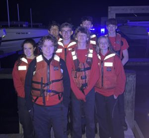 A group of eight people wearing red shirts and orange life vests are standing on a dock at night. Boats are visible in the background. Some members are smiling. The scene is dimly lit with a faint blue glow in the background.