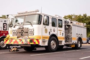 A white fire truck with yellow and red accents is parked on a paved area. It is labeled "Engine Co. 2" and equipped with various firefighting tools. Nearby, part of another fire truck with the number "3" is visible. Trees are in the background.