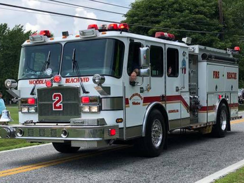 Fire truck from the Woodland Beach Volunteer Fire Department driving on a road. The truck is white with red stripes and has emergency lights on top. Trees and utility lines are visible in the background.