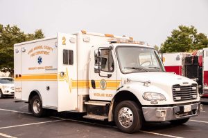 A white emergency medical services (EMS) vehicle from Anne Arundel County Fire Department, marked as unit 2, is parked in a parking lot. It has yellow stripes and a medical insignia, with trees visible in the background.
