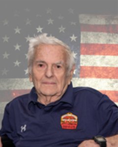 An elderly person wearing a navy blue shirt with a patch sits in front of a faded American flag backdrop. The person has white hair and a neutral expression.