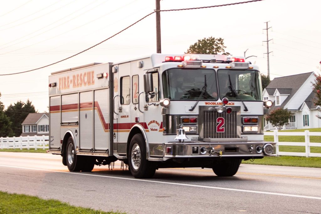 Firetruck with the number 2 and "Fire-Rescue" labeled on the side, driving on a residential road with white fences. The vehicle's lights are flashing. Houses and trees are visible in the background under a cloudy sky.