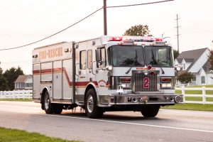 Firetruck with the number 2 and "Fire-Rescue" labeled on the side, driving on a residential road with white fences. The vehicle's lights are flashing. Houses and trees are visible in the background under a cloudy sky.
