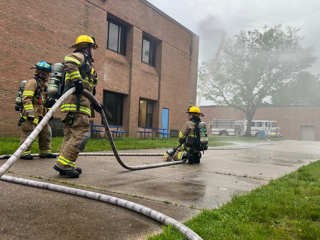 Firefighters in protective gear manage hoses outside a brick building, with smoke visible in the background. A fire truck is parked nearby. A large tree stands beside the building, partially shrouded in smoke.
