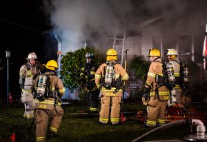 Firefighters in yellow helmets and gear gather outside a smoke-filled building at night. A ladder leans against the structure, and equipment is scattered on the grass. Thick smoke billows near the roof.