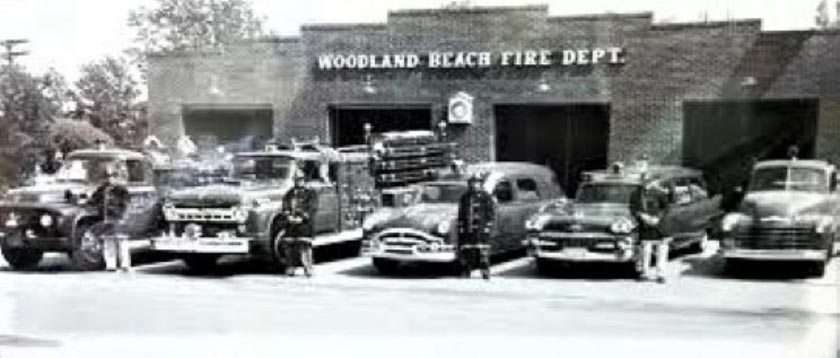 Black and white photo of the Woodland Beach Fire Dept. featuring vintage fire trucks parked in front of a brick fire station. Several firefighters in uniforms stand beside the vehicles.