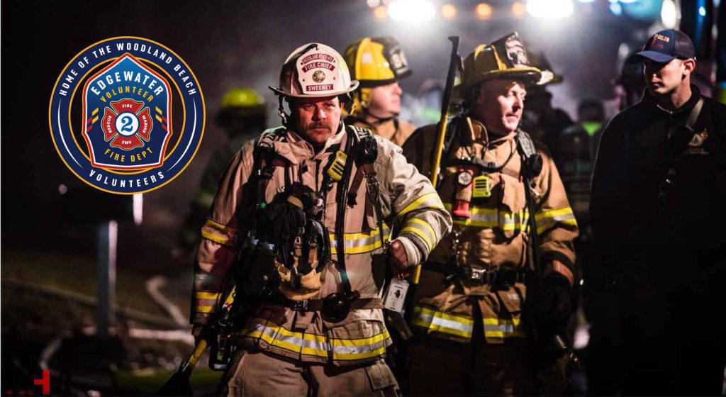 Firefighters in full gear, including helmets and reflective jackets, walk through a dimly lit scene at night. A fire department badge with "Edgewater Volunteer Fire Dept." is visible on the left. Emergency lights are visible in the background.