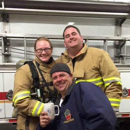 Three people stand in front of a fire truck. Two wearing firefighter gear, one holds a mug while smiling. The third in a casual jacket and hat stands in front, also smiling. They appear to be in a fire station.
