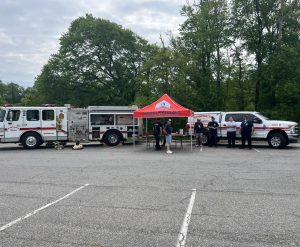 A fire department team stands beside a fire truck, an SUV, and a red canopy tent in a parking lot with trees in the background. Equipment is displayed near the vehicles, and several people are gathered under the tent.