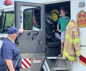 A firefighter stands by a fire truck as a smiling child wearing a red helmet and green shirt stands on the truck's step, holding a firefighter's coat with equipment visible inside the truck.