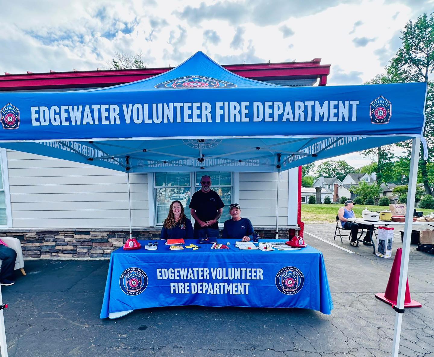 Three people sit and stand behind a table covered with Edgewater Volunteer Fire Department materials under a blue canopy tent with the department’s name, outside a building on a sunny day.