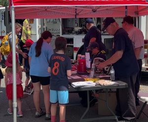 A group of people, including children and adults, gather under a red tent at an outdoor event. Some are talking to volunteers seated at a table with pamphlets, snacks, and water bottles. Firefighters and a fire truck are visible in the background.