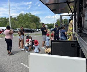 Children wearing red fire hats stand in line with adults outside a fire truck, talking to firefighters in uniform. The scene takes place in a parking lot on a sunny day with trees and parked cars in the background.