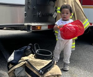 A young child stands next to a fire truck, holding a red firefighter helmet. Firefighter gear is on the ground nearby, and the child looks up with a curious expression.