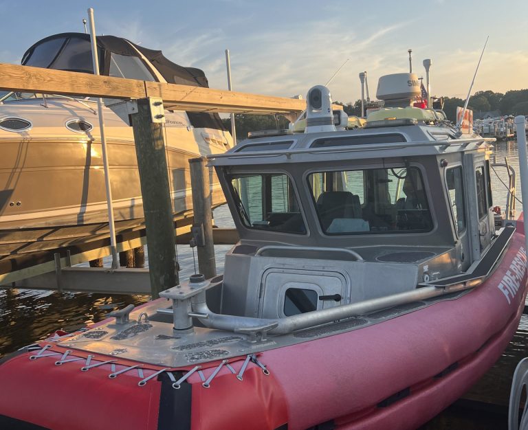 A small red and silver patrol boat is docked at a marina during sunset, with another larger boat on a lift nearby and calm water reflecting the sky.