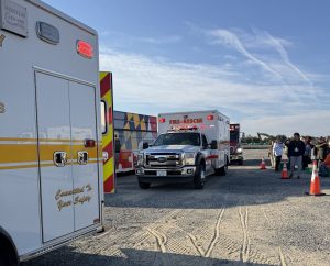 Several ambulances and emergency vehicles are parked on a gravel lot near traffic cones. A group of people stands nearby, and the sky is clear with light clouds overhead.